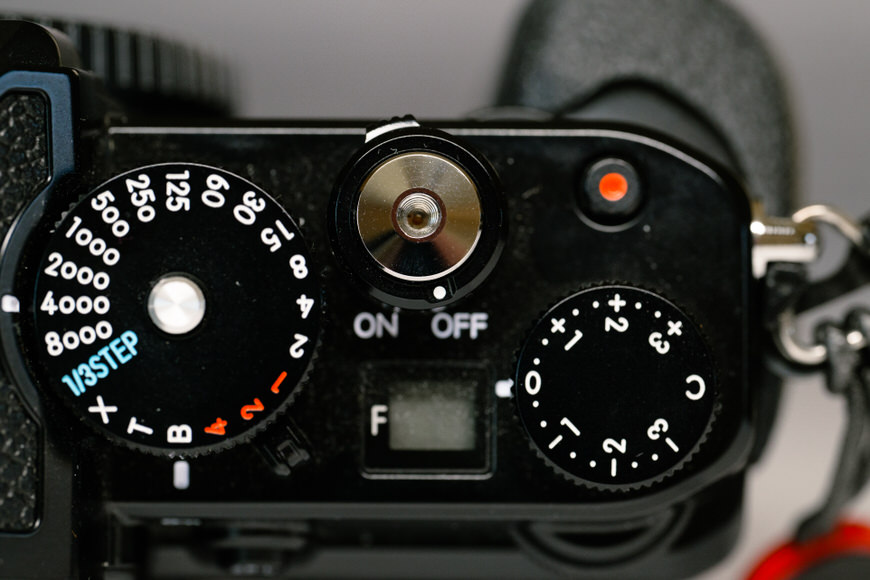 Close-up view of a camera top showing dials for shutter speed, exposure compensation, an on/off switch, and a red dot indicator.