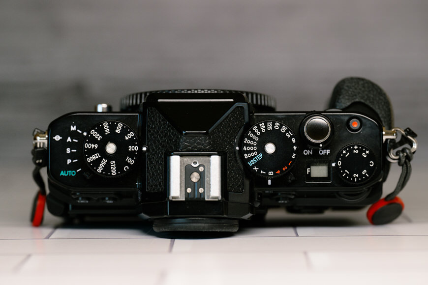 Top view of a black camera with various control dials and buttons, including ISO, shutter speed, and power switch, set against a blurred background.