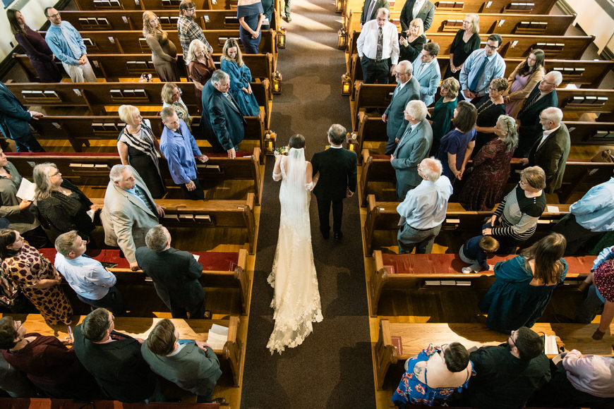 Bride walking down the aisle with an older man in a church, surrounded by standing guests.
