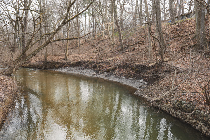 A calm, narrow river flows through a forest of bare trees on a cloudy day, with a sloped, leaf-strewn embankment on one side.