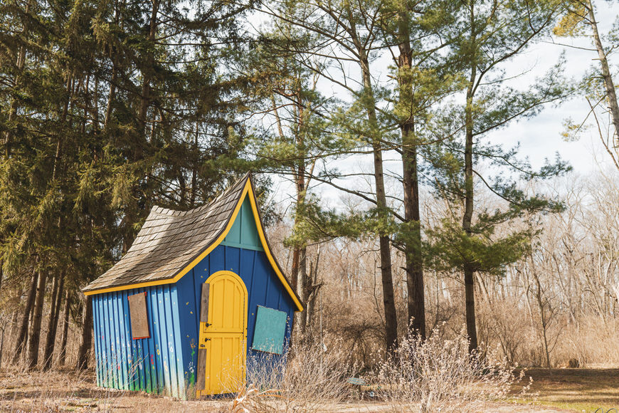 A small, colorful wooden house with a tilted roof stands among tall trees in a forested area.