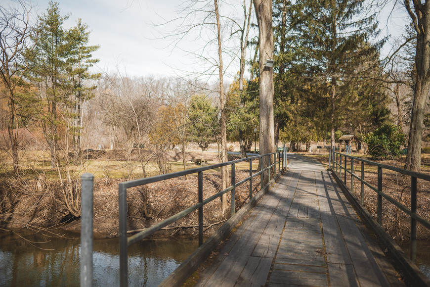 A wooden bridge with metal railings crosses over a narrow stream in a forested area. Bare trees and sparse greenery surround the scene.