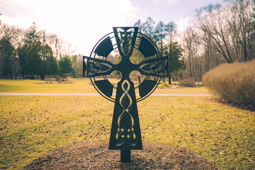 A large Celtic cross stands in a grassy park. Trees and a path are visible in the background under a partly cloudy sky.