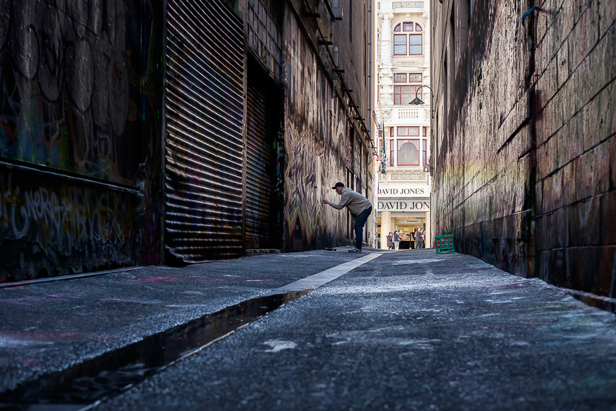A person skateboards in a narrow graffiti-filled alleyway on a sunny day with a building and a sign for "David Jones" visible in the background.