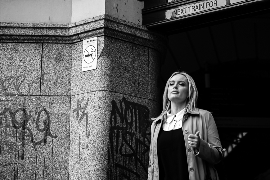 A person in a trench coat walks past a graffiti-marked wall near a "No Smoking" sign and a train station entrance.