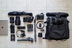 Flat lay of travel photography gear on a tiled floor, including a laptop, passport, camera lenses, cameras, chargers, memory cards, batteries, tripod, cables, and a black backpack.