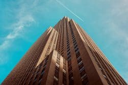 A view from below of a tall, brown skyscraper set against a bright blue sky with a contrail visible.