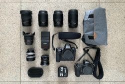 An assortment of photography equipment neatly arranged on a tiled floor, including two cameras, multiple lenses, a flash, a battery pack, and a camera bag.