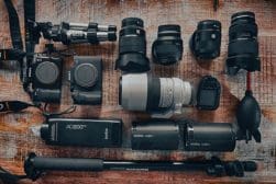 Assorted photography equipment including cameras, lenses, flashes, a monopod, and cleaning tools arranged on a wooden surface.