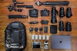 An array of camera and video equipment including cameras, lenses, tripods, a drone, a backpack, a laptop, and accessories organized on a wooden floor.