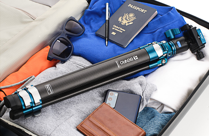 A suitcase containing a tripod, passport, folded clothes, sunglasses, a smartphone, a notebook, and a wallet.