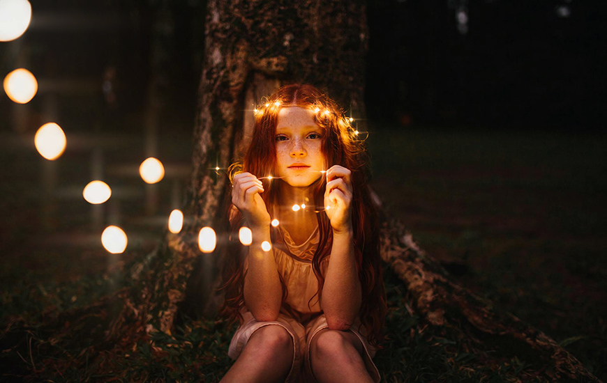 A person with long hair sits by a tree at night, holding glowing string lights with a thoughtful expression.