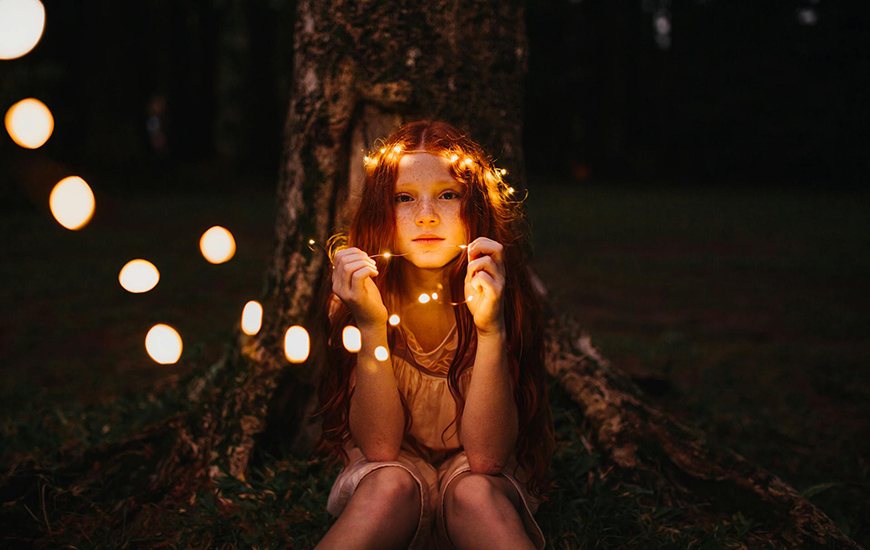 A girl with red hair sits by a tree, holding glowing fairy lights in a dimly lit forest setting.