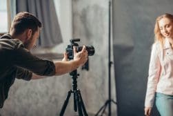 A photographer is taking pictures of a woman in a studio setting. The woman is standing in front of a grey backdrop, and the camera is mounted on a tripod.