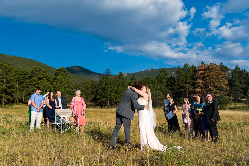 A couple kisses in a wedding ceremony outdoors, surrounded by a small group of guests, with mountains and trees in the background.