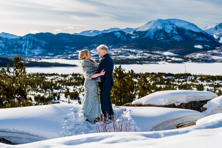 A couple in formal attire stands on a snowy landscape with mountains in the background, embracing each other under a clear sky.