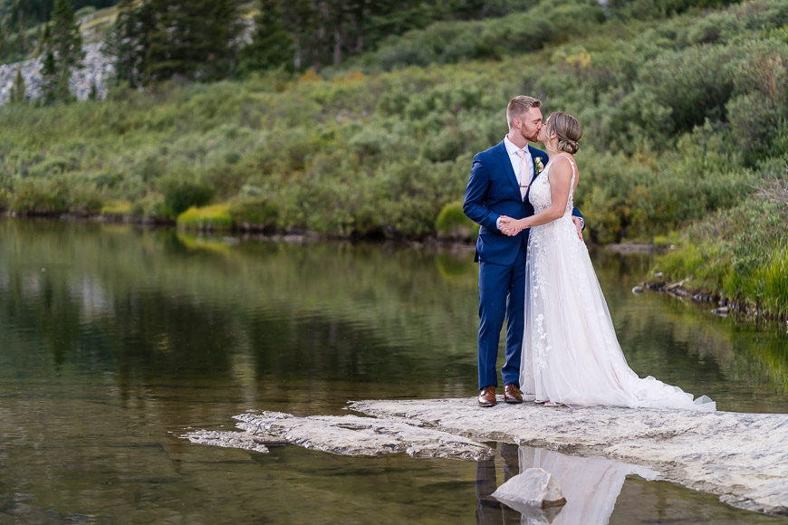 A bride and groom stand on a rock by a serene lake, surrounded by greenery. The groom wears a blue suit, and the bride wears a white dress.