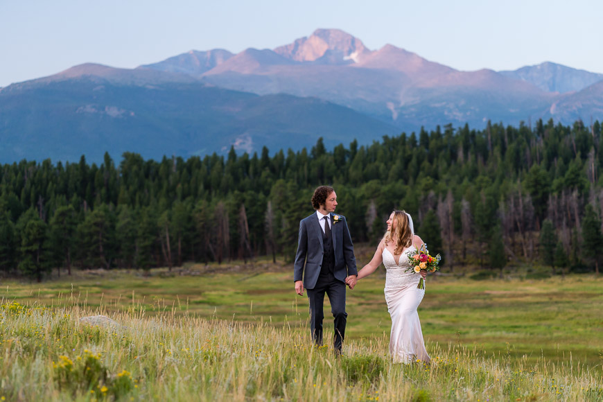 A bride and groom hold hands while walking through a grassy field with a mountain range in the background.