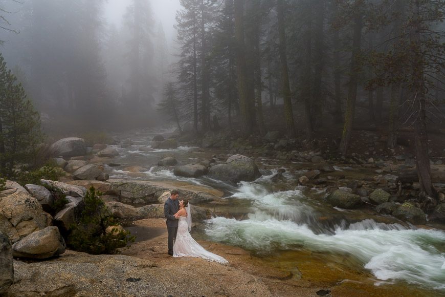 A couple embraces on a rocky riverbank in a foggy forest setting.