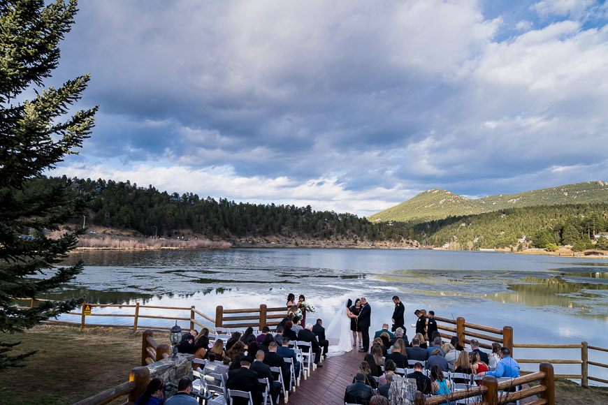 An outdoor lakeside wedding ceremony with guests seated. Mountains and trees surround the lake under a partly cloudy sky.
