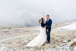 A couple in wedding attire stand on a snowy mountain landscape, smiling at each other with mist-covered peaks in the background.