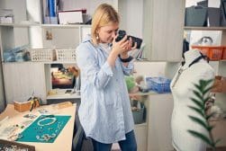 Woman in a light blue shirt takes photos of jewelry displayed on a mannequin in a craft workspace. Tools and additional jewelry items are visible on a desk beside her.