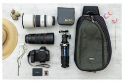 Photography gear flat lay: camera, two lenses, memory cards, mini tripod, camera bag, lens pouch, hat, and flowers on a light surface.