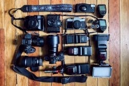 A collection of photography equipment, including various cameras, lenses, flashes, battery chargers, a light meter, and an LED light, neatly arranged on a wooden surface.