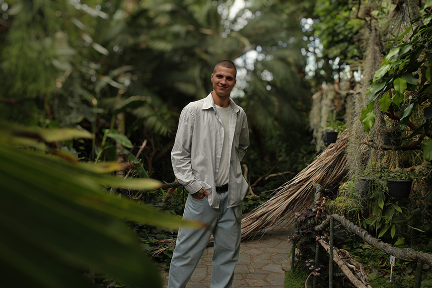 A person stands on a stone path in a lush garden, wearing a light-colored shirt and pants.