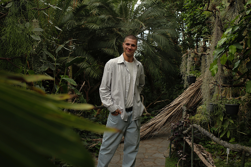 A person in a light jacket and jeans stands on a stone path surrounded by lush green foliage.