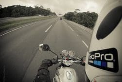Motorcyclist wearing a helmet with a GoPro sticker rides on a highway, view from their perspective, surrounded by trees and cloudy skies.