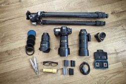 Photography equipment on wooden floor: tripod, two cameras with lenses, additional lenses, filters, multi-tool, glasses, batteries, charger with batteries, memory cards, and carrying case.