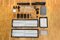 Photography equipment arranged on a wooden floor, including tripods, softboxes, a camera, lenses, wireless transmitters, light stands, power cords, and a laptop.