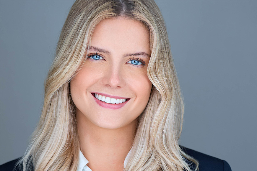 A person with long blonde hair and blue eyes smiles, wearing a dark blazer over a light shirt against a gray background.