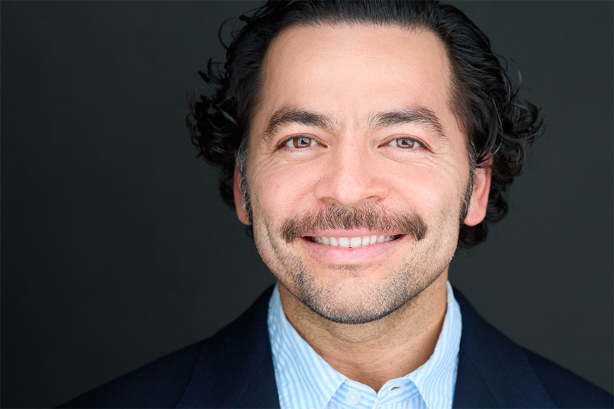 A man with curly hair and a mustache smiles, wearing a blue shirt and dark jacket against a black background.