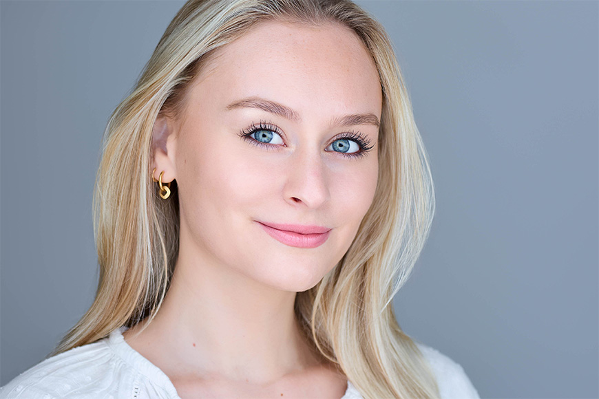 A person with long blonde hair and blue eyes is smiling slightly against a plain background. They are wearing a white top and gold hoop earrings.