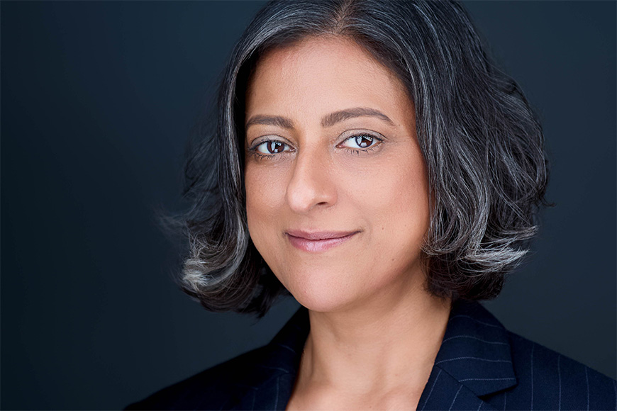 A person with shoulder-length hair and a pinstripe blazer smiles slightly against a dark background.