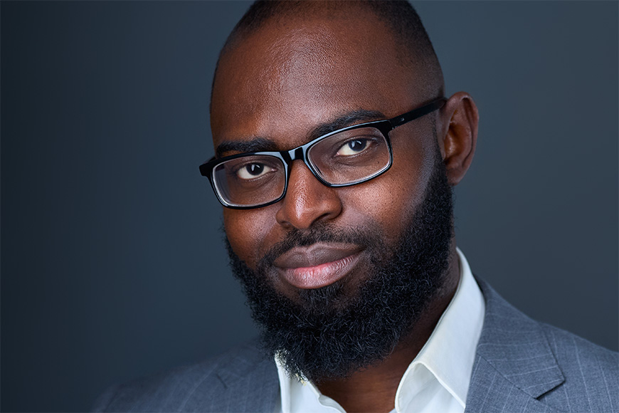 A person wearing glasses and a suit jacket is looking at the camera against a dark background.