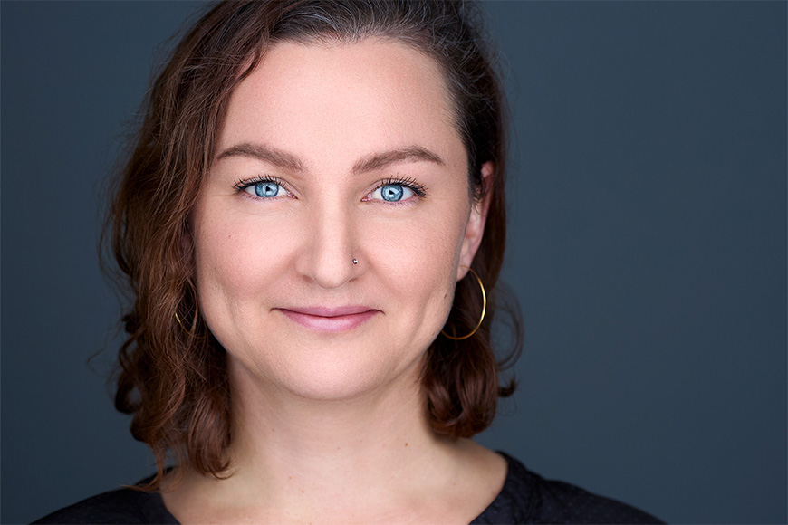 Person with short brown hair and blue eyes, wearing hoop earrings, smiles against a plain dark background.