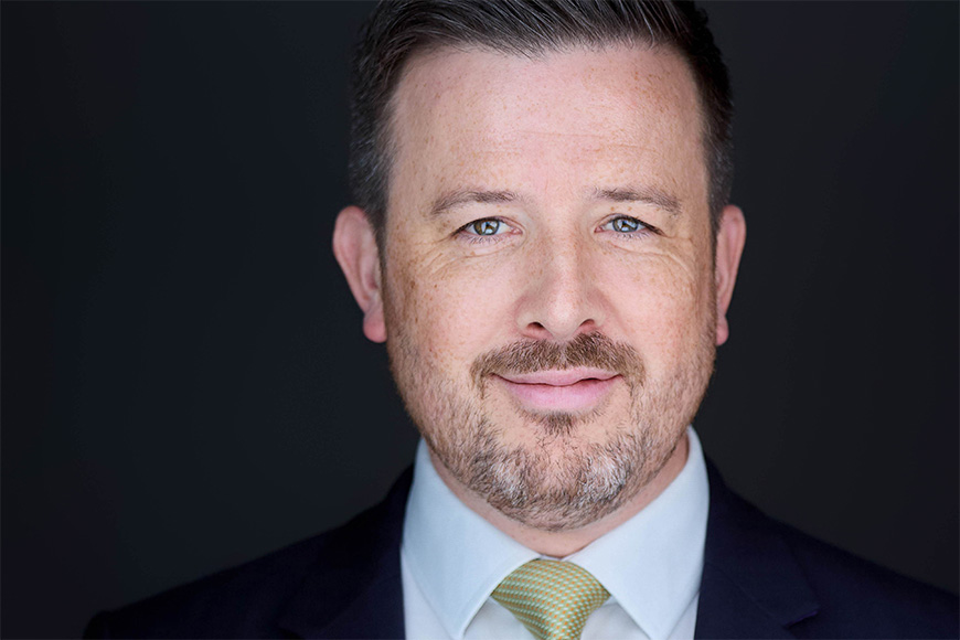 Man with short hair, beard, and mustache wearing a suit and yellow tie against a plain dark background.