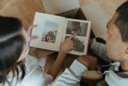 Two people sit on the floor looking at a photo album with pictures of them and a child. One person points at a photo while the other flips the page. A cardboard box is nearby.