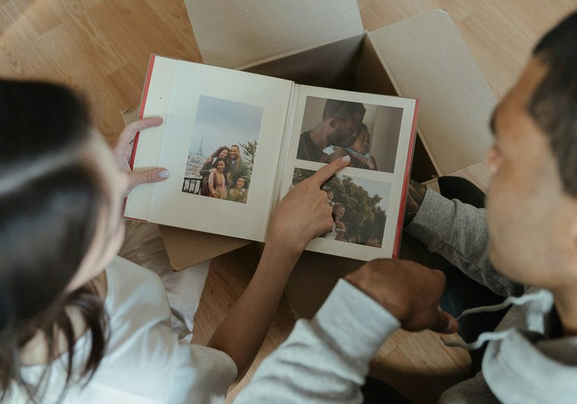 Two people sit on the floor looking at a photo album with pictures of them and a child. One person points at a photo while the other flips the page. A cardboard box is nearby. Two people sit on the floor looking at a photo album with pictures of them and a child. One person points at a photo while the other flips the page. A cardboard box is nearby.