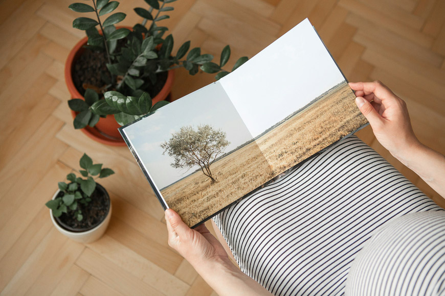 Person in striped clothing holding an open book displaying a photo of a solitary tree in a field, with potted plants on the wooden floor nearby.