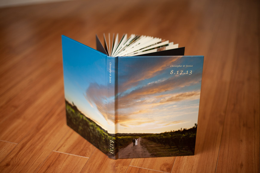 An open photo book stands on a wooden floor, displaying a scenic cover with a couple walking under a sunset. The date "8.17.13" and the names "Christopher & Jessica" are written on the cover.