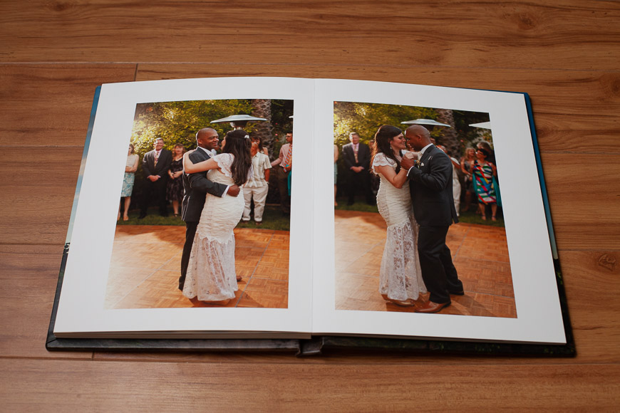 A photo album lies open on a wooden floor, displaying two photos of a bride and groom dancing outdoors during their wedding, with guests watching in the background.