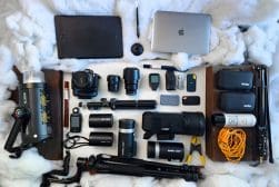 Photography and videography equipment laid out on a white surface, including a laptop, graphics tablet, camera, lenses, lighting gear, tripods, and various accessories.