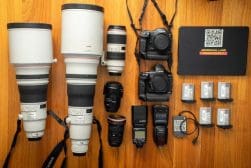 A collection of photography equipment including cameras, lenses, battery packs, flash units, memory cards, and a laptop with a label that reads "Aboriginal Land Tread Lightly," neatly arranged on a wooden surface.