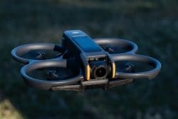 A black quadcopter drone with a central camera is flying outdoors. The drone has four propellers, each within its own circular shielding.