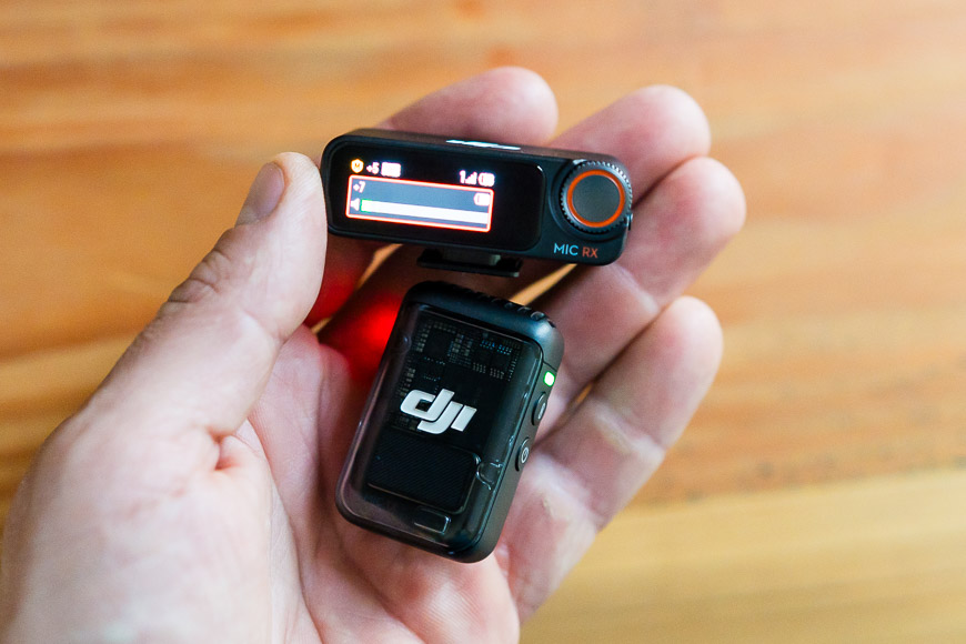 A hand holds a DJI wireless microphone receiver and transmitter against a wooden background.