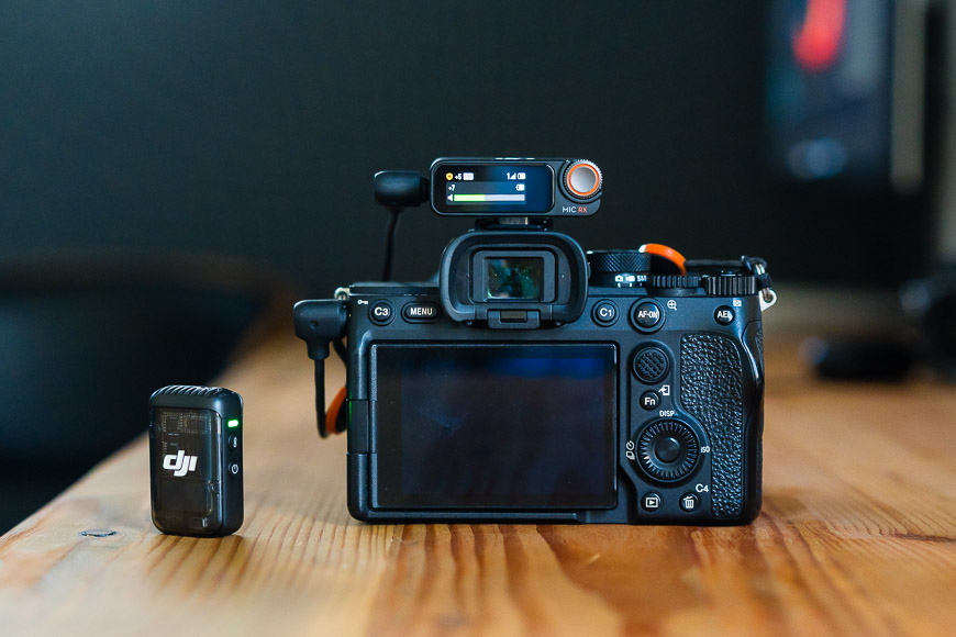 A digital camera with an external device mounted on top is placed on a wooden surface. Another small electronic device is positioned next to the camera.